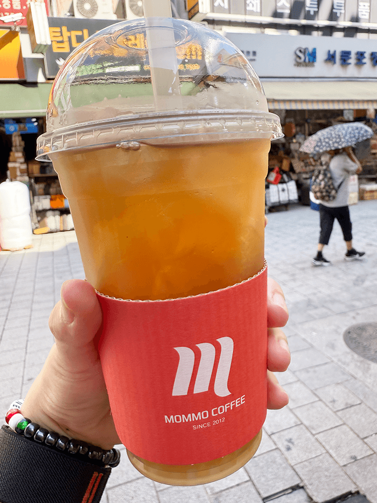 A blown tea in the plastic cup. My left hand holding it. The orange-red paper sleeve is put around the bottom of the cup