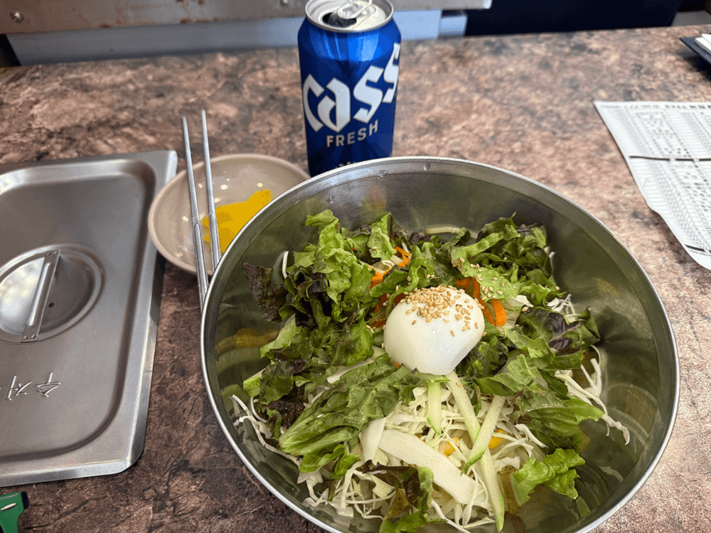 shredded veggies and half of the boiled egg are in the stainless bowl. under the veggies, slightly noodles and red sauce are showing. There are three slices of yellow pickled radish and a can of Cass beer on the table too.