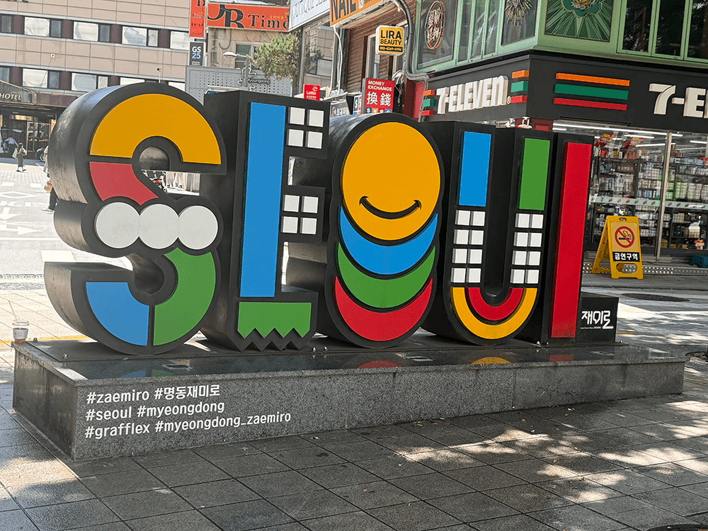 A colorful object of the word SEOUL on the street. It was made by orange, red, white, green, blue and black panel, put on the stone base. These is a 7-eleven on the back.