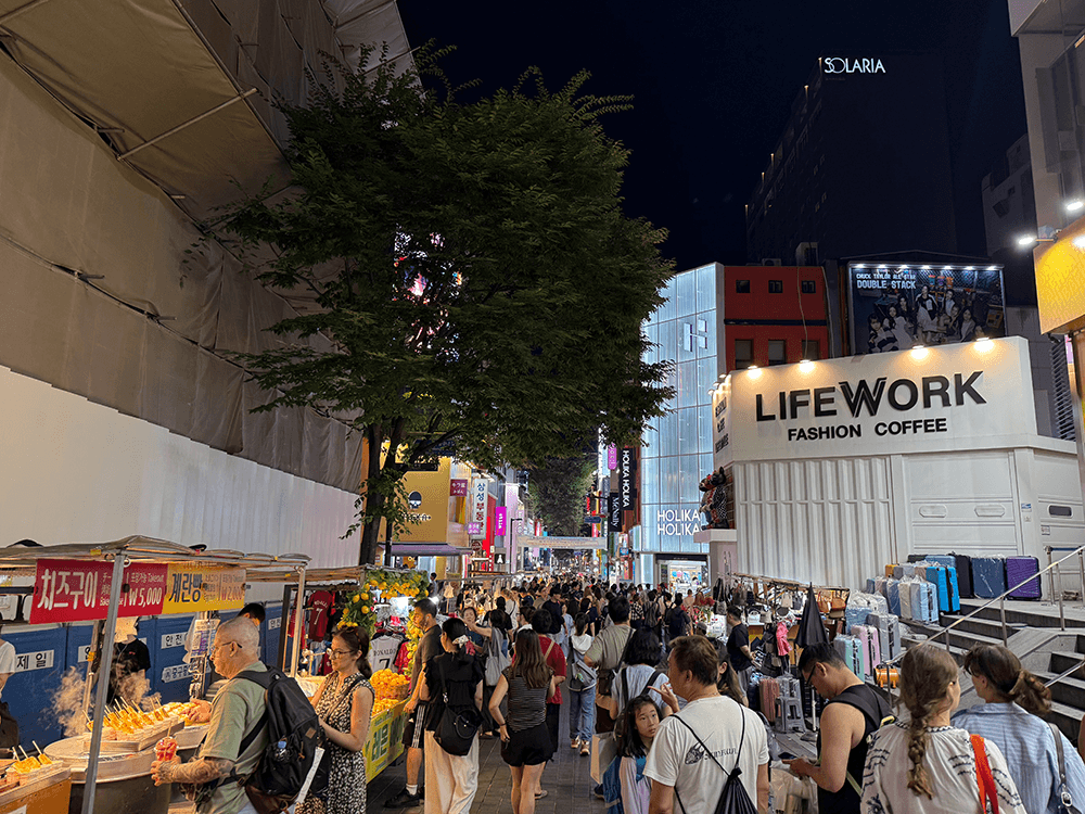 The night street there are a lot of people. There are some food stands on the left side. Both sides of the street, there are many of buildings.