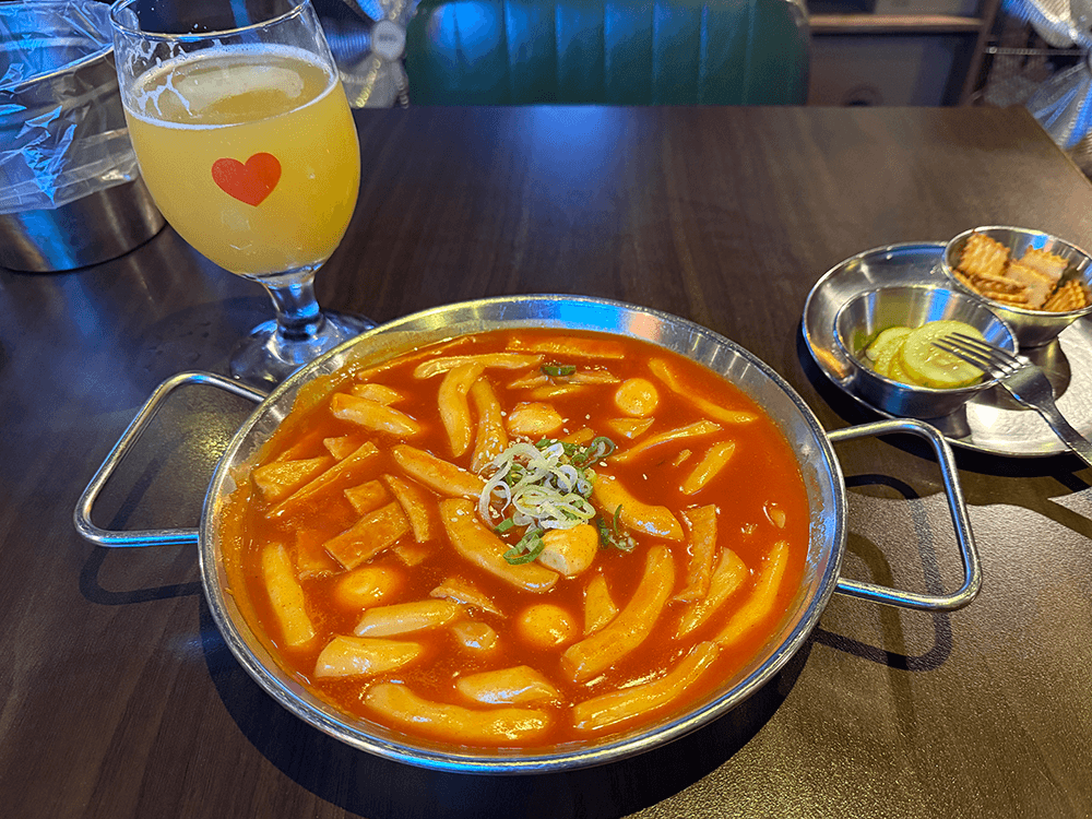 Tteokbokki, a glass of hazy beer, pickles and crackers on the table. tteokbokki is saucy, there are cylinder shaped rice cakes, the fishcakes that cut into bitesize pieces, quill eggs. A red heart is printed on beer glass.