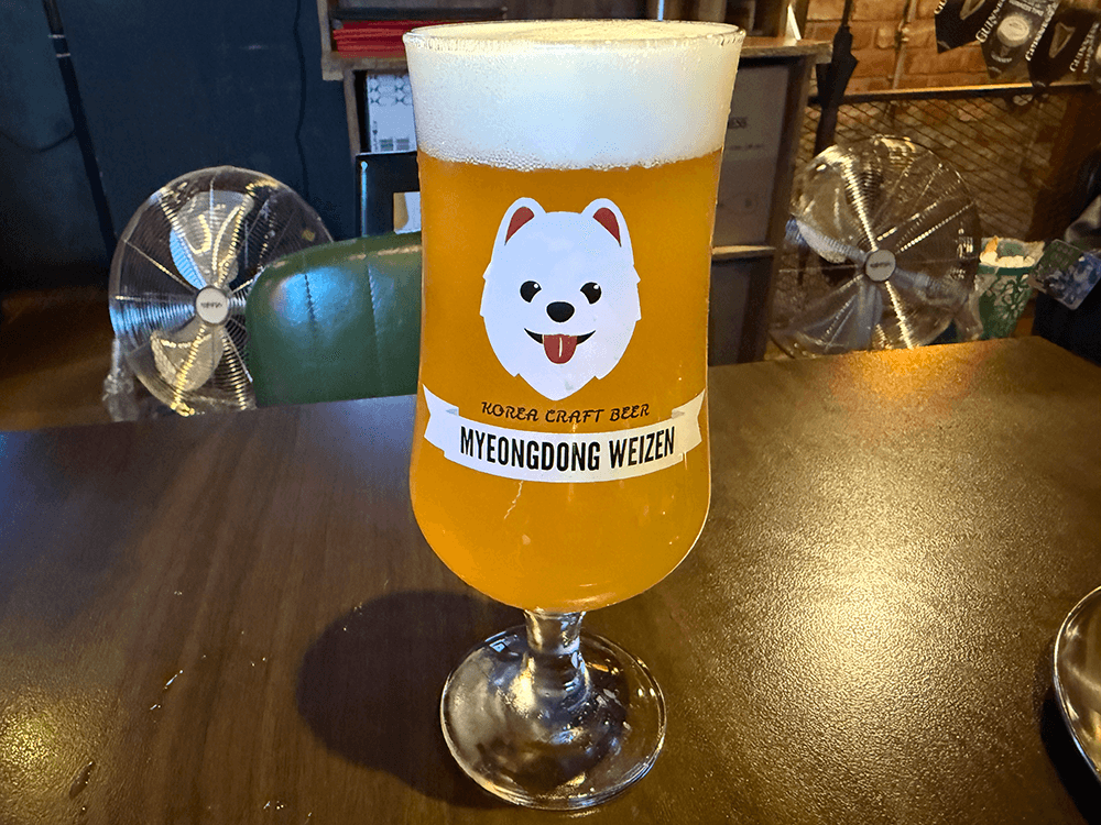 A glass of Weizen beer. A white long hair dog head that showing their tongue. It's written Korea craft beer, Myeondong Weizen.