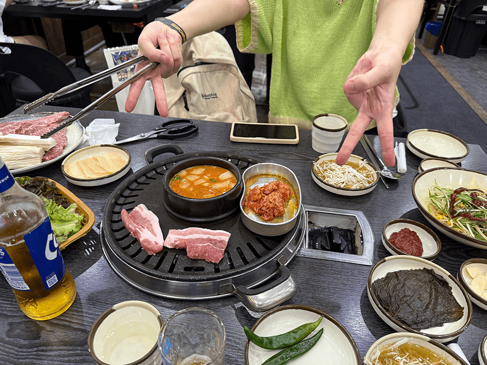 A table of Korean BBQ restaurant, there is the grill on the center. Sliced porks, Kimchi with sesame oil in the kidney been shaped stainless pan, and doenjang jjigae in the earthenware pot are on the grill. My friend wearing green knit tops making V by both hands.