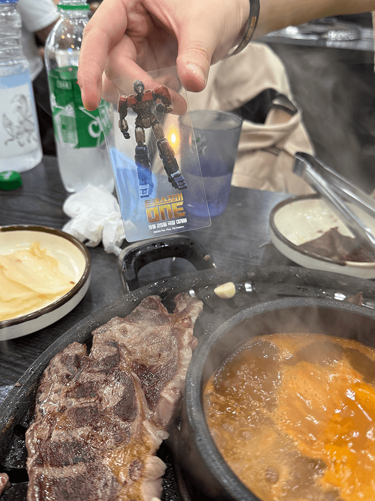 There is a big slice of grilled beef on the plate, my friend is showing TF ONE Orion Pax acrylic card.